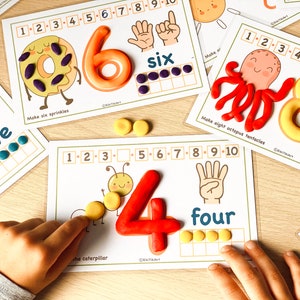 May include: A set of printable learning cards for kids featuring colorful play dough numbers and counting activities. The cards include a donut with sprinkles, an octopus with tentacles, a caterpillar, and a hand with fingers. The text on the cards includes the numbers 1 through 10, the words "six", "five", "four", and instructions to "Make six sprinkles", "Make eight octopus tentacles", and "Make the rainbow".