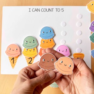 May include: A learning activity featuring colorful ice cream cone cutouts with numbers 1 and 2, and the text "I CAN COUNT TO 5". The ice cream scoops are various colors, including pink, green, and blue. The activity is designed for early childhood education.