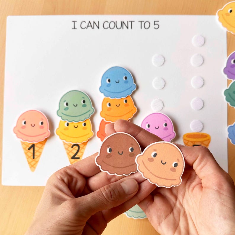 May include: A learning activity featuring colorful ice cream cone cutouts with numbers 1 and 2, and the text "I CAN COUNT TO 5". The ice cream scoops are various colors, including pink, green, and blue. The activity is designed for early childhood education.
