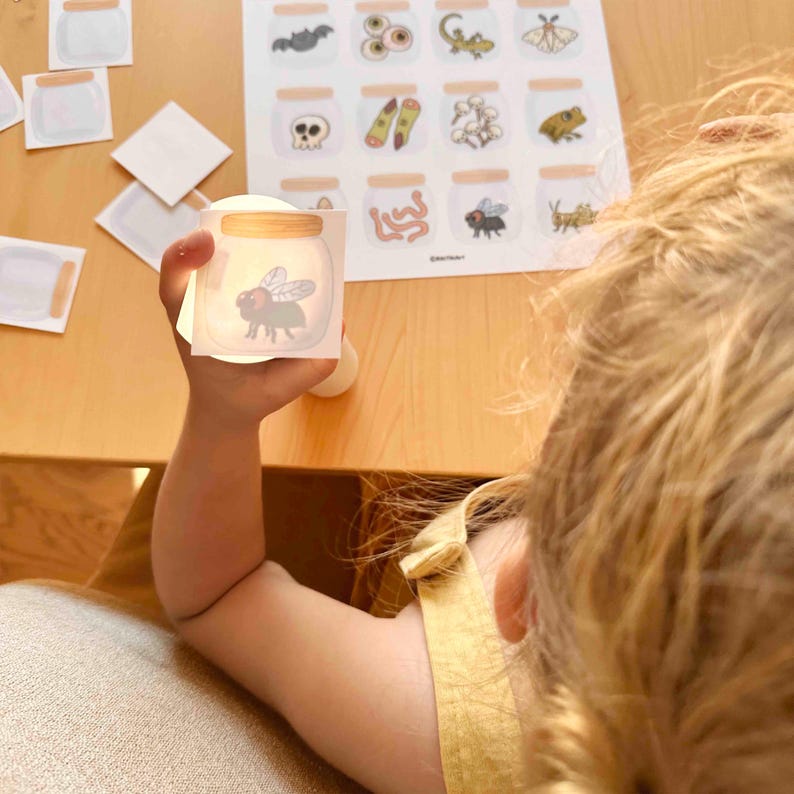 Puede incluir: Una tarjeta cuadrada que muestra una mosca de dibujos animados dentro de un frasco de vidrio con tapa color canela. La tarjeta se sostiene frente a una hoja que contiene una cuadr&iacute;cula de ilustraciones de frascos similares, incluyendo un murci&eacute;lago, ojos, lagarto, polilla, calavera, dedos, algod&oacute;n, rana, gusanos y un saltamontes.