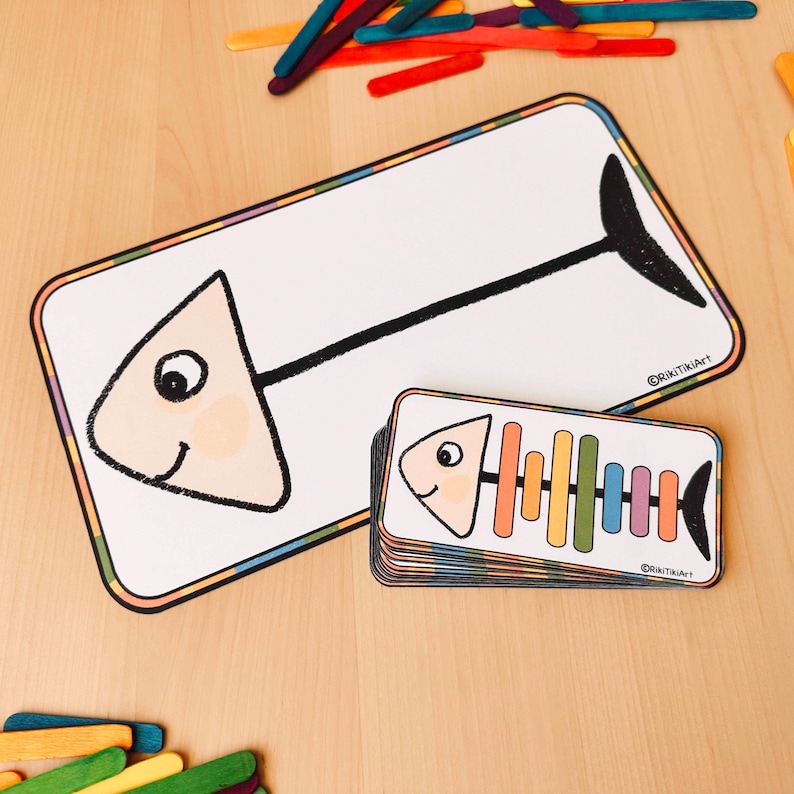 May include: A black and white outline of a fish with a colourful rainbow skeleton. The fish is on a white card with a black outline and a colourful border. The card is on a wooden surface.