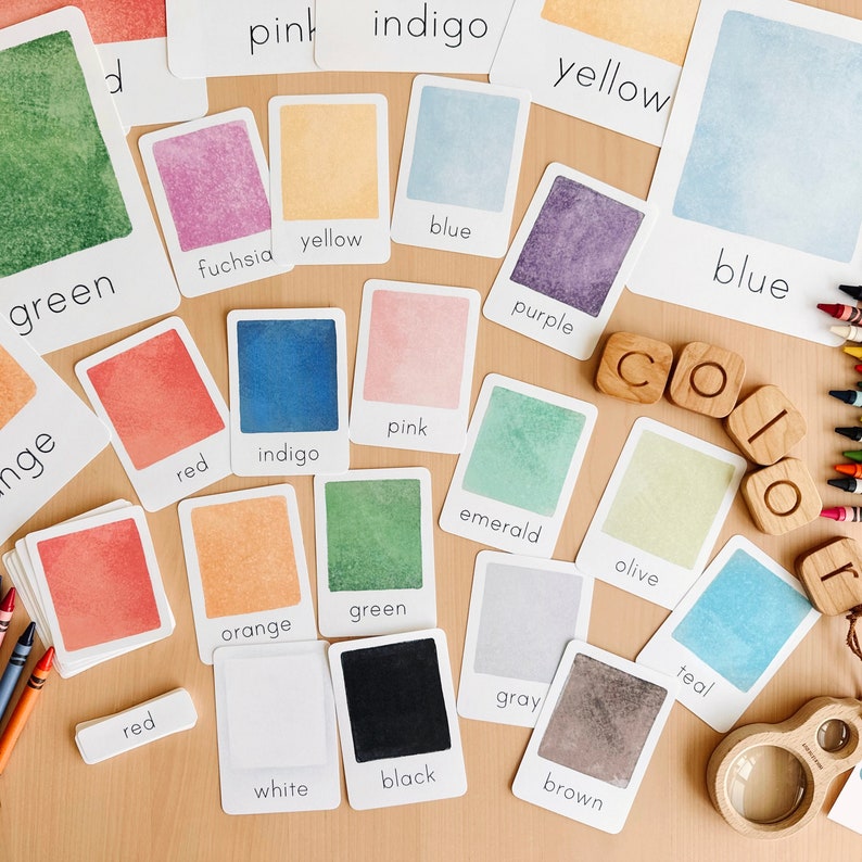 May include: A set of color cards with watercolor painted swatches and the corresponding color names written below each swatch. The colors include red, orange, yellow, green, blue, purple, pink, brown, gray, teal, olive, black, white, fuchsia, indigo, and emerald. The word "COLOR" is spelled out with wooden letter tiles.