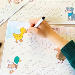 May include: A child's hand traces dotted lines on a white sheet of paper with a black marker. The paper has illustrations of a duck, a goose, and a turkey. The paper also has dotted lines in the shape of zigzags, waves, and squares. The paper has the text "Tracing" at the top.