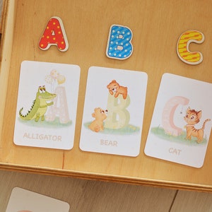 May include: A wooden box with three alphabet cards featuring an alligator, a bear, and a cat. The cards are labeled with the letters A, B, and C. The box also has three wooden letters, A, B, and C, on top of the cards.