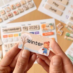 以下が含まれることがあります： 日付、月、季節の交換可能なタイルが付いた木製のカレンダーセット。 「Winter」の文字と雪だるまのイラストが描かれたカードがカレンダーの前に掲げられています。 他のカレンダーコンポーネントが背景に見えます。