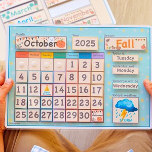 May include: A wooden calendar with a blue frame and a light blue background. The calendar is for October 2025 and shows the days of the week, the date, and the season, which is Fall. The calendar also includes a weather icon for a storm.