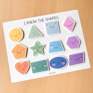 Pu&ograve; includere: Una lavagna bianca con adesivi di forme colorate e sorridenti. Le forme includono un cerchio, un triangolo, un quadrato, un rombo, un pentagono, una stella, un trapezio, un esagono, un cuore, un rettangolo e un ovale. Il testo dice "I KNOW THE SHAPES".