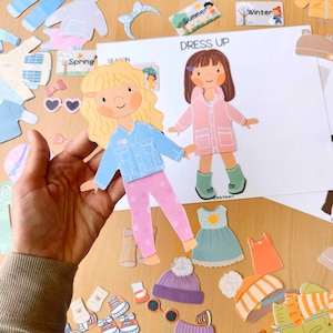 May include: A hand holds a paper doll wearing a blue denim jacket and pink polka dot pants. The doll is part of a "Dress Up" set with various clothing items and accessories for different seasons, including a pink raincoat, hats, and sunglasses.