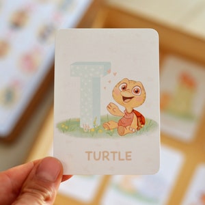 May include: A white card with a light blue letter T and a cartoon turtle with a red shell. The turtle is smiling and waving. The card says "TURTLE" in pink letters.