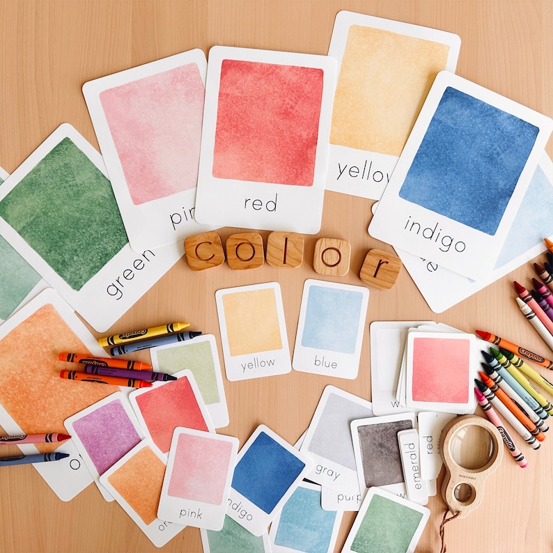 May include: A set of color cards with watercolor painted swatches and the color name printed below each swatch. The colors include green, pink, red, yellow, indigo, blue, gray, and orange. The cards are arranged on a wooden surface with wooden letter blocks spelling out the word "color".