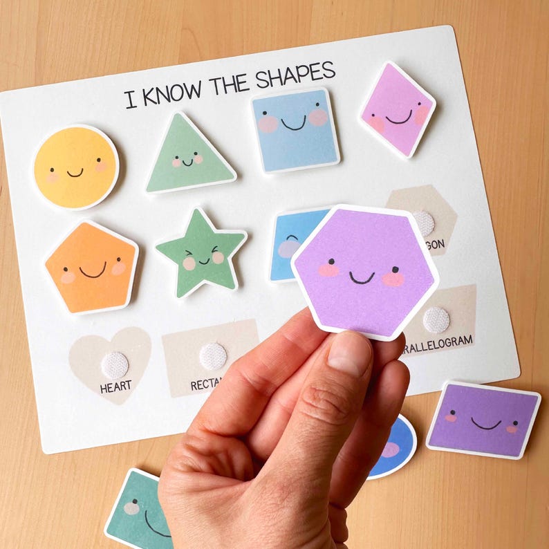 Pu&ograve; includere: Una lavagna bianca con la scritta "I KNOW THE SHAPES" e ritagli di forme colorate con facce sorridenti. Le forme includono un cerchio, un triangolo, un quadrato, un rombo, un pentagono, una stella, un esagono e un parallelogramma. Una mano tiene in mano un ritaglio esagonale viola.
