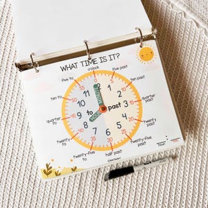 May include: A spiral-bound educational chart with a clock face, designed to teach time-telling. The clock face is yellow with black numbers and labels like "quarter past" and "ten to." The text "WHAT TIME IS IT?" is at the top. A black marker rests below.
