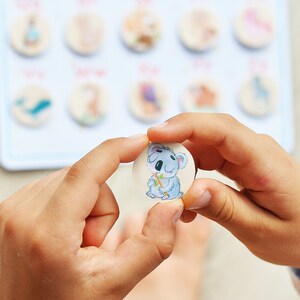 May include: A child's hand holds a round wooden token with a cartoon koala bear illustration. The koala is gray and white with a brown nose and is holding a green stick.