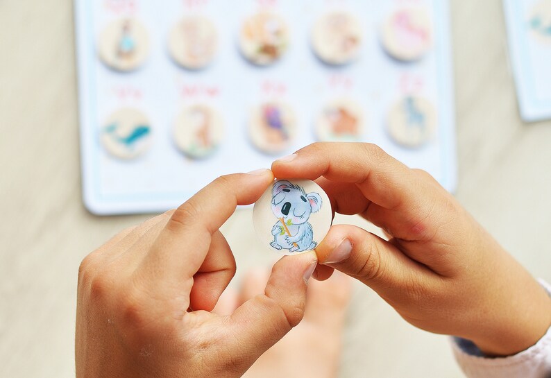 May include: A child's hand holds a round wooden token with a cartoon koala bear illustration. The koala is gray and white with a brown nose and is holding a green stick.