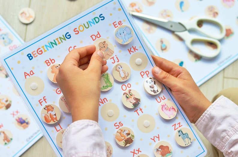 May include: A colorful learning chart with the title "BEGINNING SOUNDS" and a grid of animal illustrations representing different letters of the alphabet. A child's hands are shown placing a letter tile on the chart.