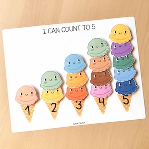 May include: A white educational counting board with the text "I CAN COUNT TO 5". The board features illustrated ice cream cones with smiling faces, each topped with a different colored scoop, numbered from one to five.
