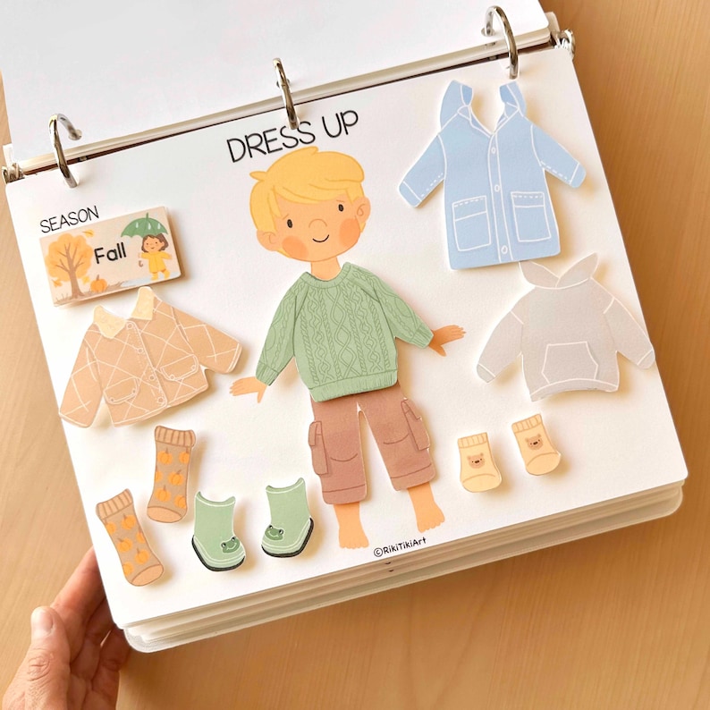 May include: A children's "Dress Up" activity book featuring a cartoon boy and various fall clothing items. Includes a green sweater, brown pants, socks, boots, a light blue raincoat, and a gray hoodie. The word "Fall" is also visible.