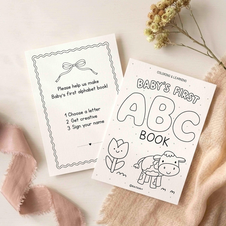 May include: Two white paper cards with black line art. One card features a bow, a wavy border, and text: Please help us make Baby's first alphabet book! 1 Choose a letter 2 Get creative 3 Sign your name. The other card reads: BABY'S FIRST ABC BOOK.