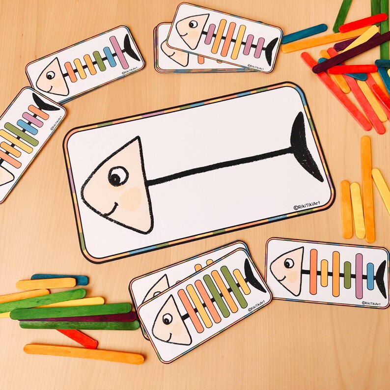 May include: A set of colourful fish-shaped cards with a blank fish card and a set of colourful wooden sticks. The cards have a black outline and a white background. The fish have a black eye and a black mouth. The sticks are arranged in a rainbow pattern. The cards are labelled with the copyright symbol and the text "@knitukart".