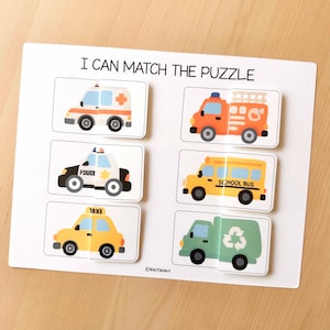 May include: A white puzzle board with the text "I CAN MATCH THE PUZZLE" at the top. The puzzle features six vehicle images: an ambulance, fire truck, police car, school bus, taxi, and a recycling truck. Each vehicle is a separate puzzle piece.