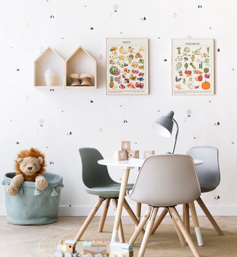 May include: A white table with four chairs, two gray and two green, surrounded by a light brown rug. A stuffed lion sits in a blue basket. A lamp with a gray shade sits on the table. The table has a set of wooden blocks with letters and numbers on them. Two framed posters hang on the wall, one titled "Fruits" and the other titled "Vegetables".
