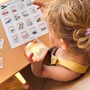 Puede incluir: Una hoja de actividades Witch's Potion con una cuadr&iacute;cula de frascos ilustrados que contienen varios elementos como huesos, hongos e insectos, junto a una peque&ntilde;a tarjeta cuadrada que muestra un saltamontes en un frasco, sostenida para iluminar el gr&aacute;fico.