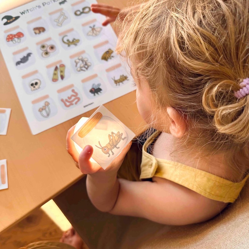 Puede incluir: Una hoja de actividades Witch's Potion con una cuadr&iacute;cula de frascos ilustrados que contienen varios elementos como huesos, hongos e insectos, junto a una peque&ntilde;a tarjeta cuadrada que muestra un saltamontes en un frasco, sostenida para iluminar el gr&aacute;fico.