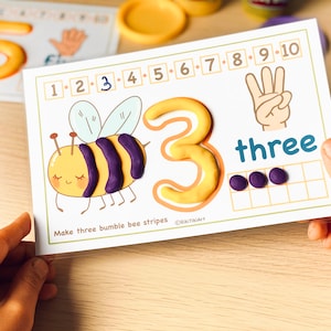 May include: A printable learning card with a yellow number 3, the word "three" and a cartoon bee with three purple stripes. The card has a grid with three purple circles in the first row. The text "Make three bumblebee stripes" is at the bottom of the card.