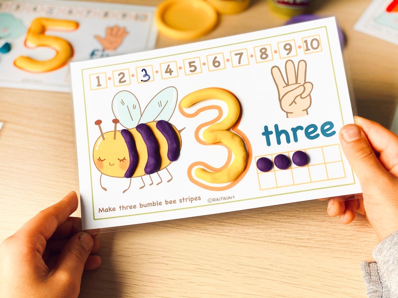 May include: A printable learning card with a yellow number 3, the word "three" and a cartoon bee with three purple stripes. The card has a grid with three purple circles in the first row. The text "Make three bumblebee stripes" is at the bottom of the card.