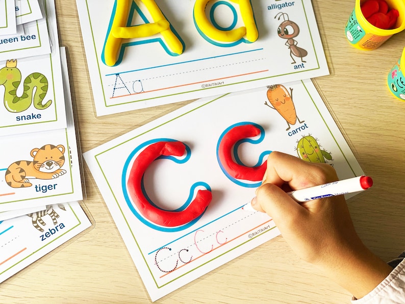 May include: A child's hand traces the letter 'C' with a marker on a white card with a blue outline. The letter 'C' is made of red playdough. The card also has a picture of a carrot and a cactus.