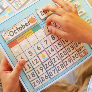 May include: A blue and white calendar with a colorful October month tab. The calendar has a grid of numbers from 1 to 30, with a small birthday cake icon on the 14th. The calendar is being used by a child to learn about the days of the month.