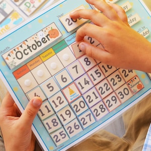 May include: A blue and white calendar with a colorful October month tab. The calendar has a grid of numbers from 1 to 30, with a small birthday cake icon on the 14th. The calendar is being used by a child to learn about the days of the month.