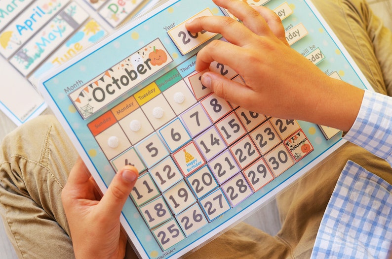 May include: A blue and white calendar with a colorful October month tab. The calendar has a grid of numbers from 1 to 30, with a small birthday cake icon on the 14th. The calendar is being used by a child to learn about the days of the month.
