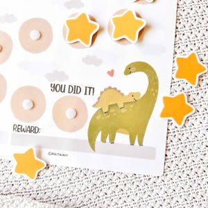 May include: A reward chart with a dinosaur theme. The chart features a large green dinosaur with a smaller yellow dinosaur on its back, along with star-shaped stickers. Text on the chart reads "YOU DID IT!" and "REWARD:"