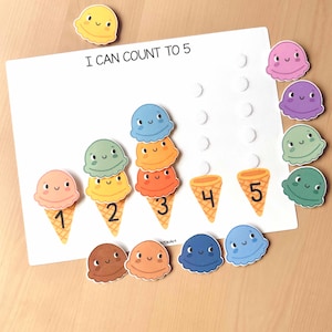 May include: A white educational board with the text "I CAN COUNT TO 5" features colorful ice cream cone illustrations. The cones are numbered 1-5, with matching ice cream scoops. Additional ice cream scoops in various colors are scattered around the board.