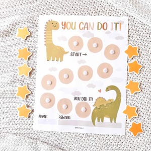 May include: A reward chart with the text "YOU CAN DO IT!" and "YOU DID IT!" featuring dinosaur illustrations. The chart has spaces for rewards and is surrounded by star-shaped stickers. The chart is on a white background.