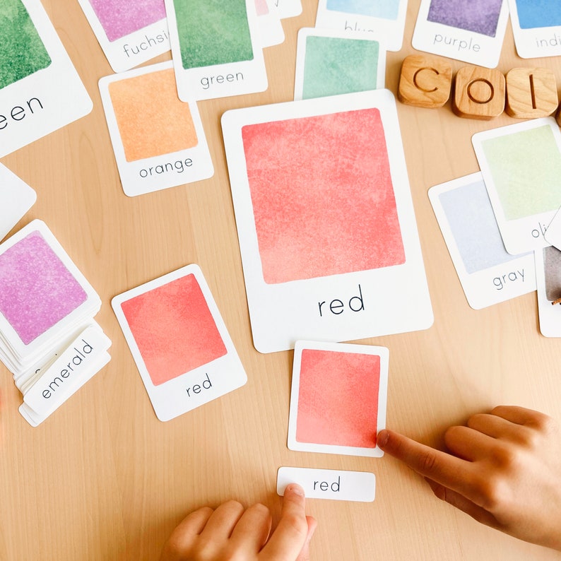 May include: A child's hand points to a red color card with the word "red" written on it. The card is part of a set of color cards with watercolor-like backgrounds. The cards are laid out on a wooden surface.