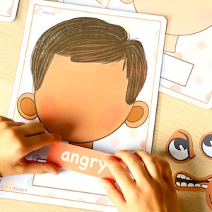 May include: A child's hand places a card with the word "angry" on a matching game board. The board features a cartoon face with brown hair and a blank expression. The game is designed to help children learn about different emotions.