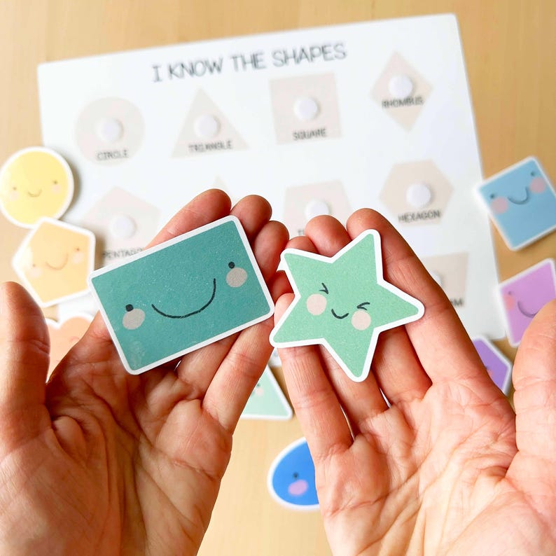 Pu&ograve; includere: Un giocattolo educativo con una lavagna bianca con la scritta "I KNOW THE SHAPES" e ritagli di forme. Due mani tengono un rettangolo verde e una stella verde, entrambi con facce sorridenti. Altre forme sono sparse.