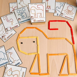 Pu&ograve; includere: Un set di bastoncini di legno colorati disposti per formare la forma di un elefante. I bastoncini sono disposti in un motivo rosso, arancione e giallo. Ci sono anche diverse carte con i contorni degli animali e il testo "Posso fare un elefante", "Posso fare un'anatra", "Posso fare un fenicottero", "Posso fare una scimmia", "Posso fare una balena", "Posso fare un polpo", "Posso fare un koala", e "Posso fare un pinguino".