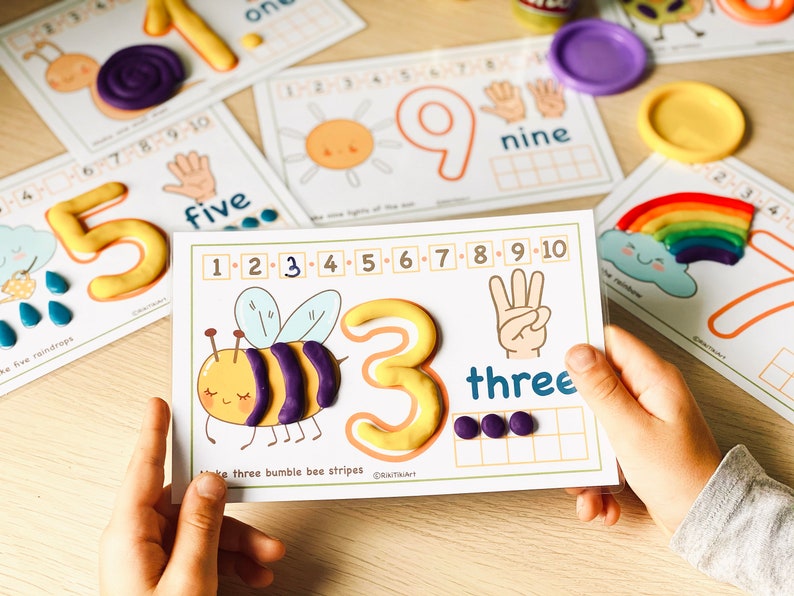 May include: A child's hand holds a card with a bee and the number three. The card has instructions to make three bumble bee stripes. There are three purple playdough stripes on the bee. The number three is made of yellow playdough. There are three purple playdough circles in a row on the card.