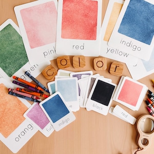 May include: A set of color learning cards with watercolor painted swatches and the color name printed below each. The colors include green, pink, red, yellow, indigo, orange, and blue. There are also wooden letter blocks that spell out the word "color" and crayons in various colors.