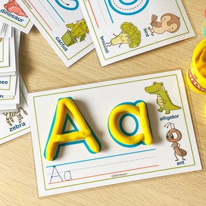 May include: A yellow play dough letter A and a yellow play dough letter a on a white card with a blue border. The card has a picture of an alligator and an ant. The text on the card says "alligator" and "ant".