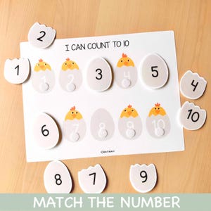 Peut inclure: Jeu éducatif de comptage avec un tableau blanc affichant "I CAN COUNT TO 10" et "MATCH THE NUMBER". Le tableau présente des formes d'œufs numérotées de 1 à 10. Des coquilles d'œufs numérotées sont éparpillées.