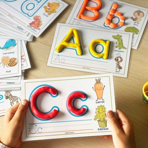 May include: A child's hand holds a white card with a blue outline around the letter C. The letter C is made of red playdough. The card has illustrations of a carrot and a cactus with the words "carrot" and "cactus" written below them. The card also has a line for tracing the letter C.