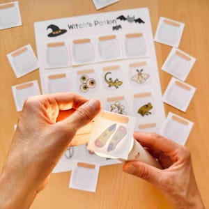 Puede incluir: Una hoja de actividades Witch's Potion con una cuadr&iacute;cula de frascos de vidrio vac&iacute;os e ilustraciones de criaturas. Una peque&ntilde;a tarjeta cuadrada que muestra dos dedos en un frasco es iluminada por una luz, con tarjetas adicionales dispersas.