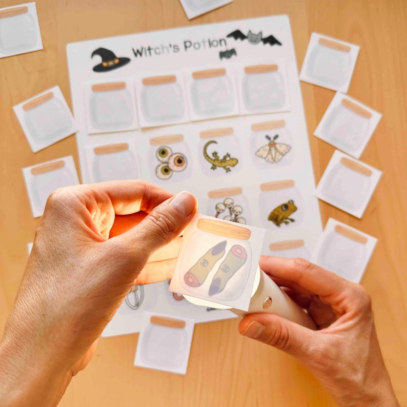 Puede incluir: Una hoja de actividades Witch's Potion con una cuadr&iacute;cula de frascos de vidrio vac&iacute;os e ilustraciones de criaturas. Una peque&ntilde;a tarjeta cuadrada que muestra dos dedos en un frasco es iluminada por una luz, con tarjetas adicionales dispersas.