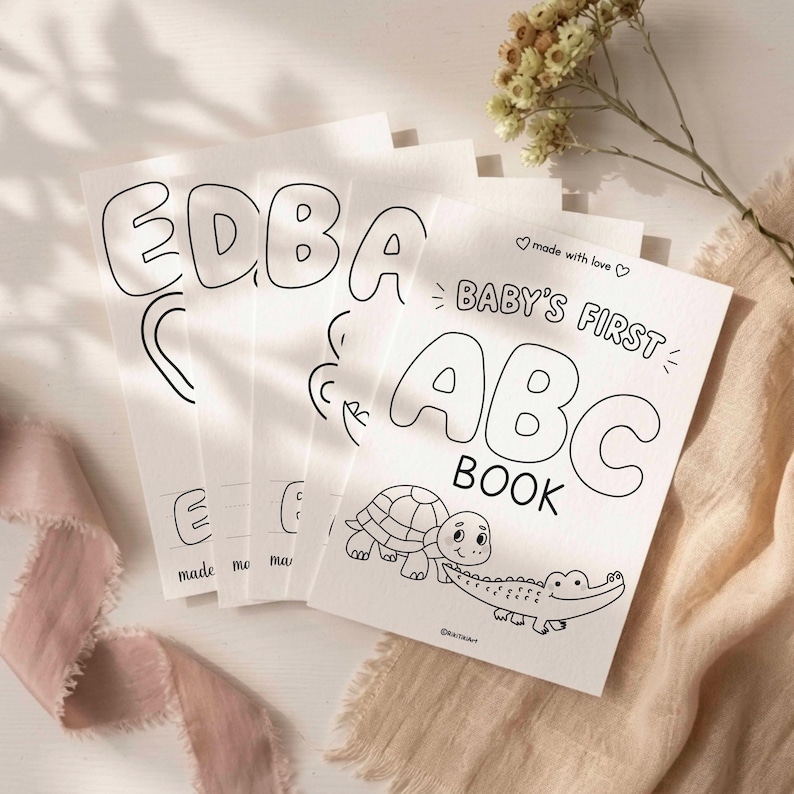 May include: A stack of white paper coloring pages titled Baby's First ABC Book, featuring black line art of a turtle and a crocodile on the cover. The pages include large bubble letters and the text made with love at the top and bottom.