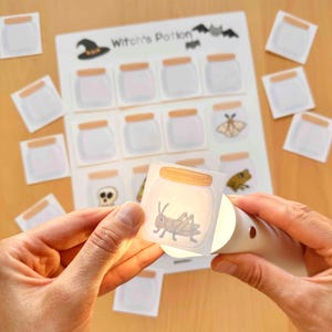 Puede incluir: Una hoja de actividades Witch's Potion con una cuadr&iacute;cula de frascos de vidrio vac&iacute;os con tapas color canela, acompa&ntilde;ada de tarjetas cuadradas separadas que representan un saltamontes, una calavera, una polilla y una rana, iluminadas por luz.