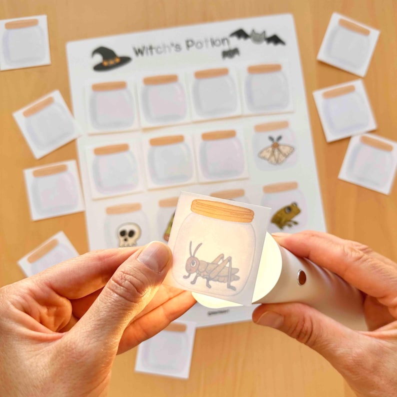 Puede incluir: Una hoja de actividades Witch's Potion con una cuadr&iacute;cula de frascos de vidrio vac&iacute;os con tapas color canela, acompa&ntilde;ada de tarjetas cuadradas separadas que representan un saltamontes, una calavera, una polilla y una rana, iluminadas por luz.
